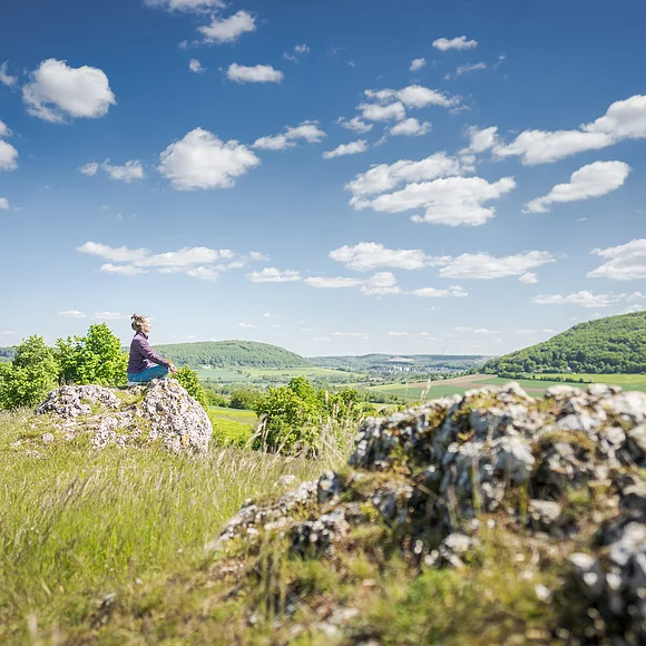 Frau sitzt auf einem Stein in Yogapose im Vordergrund ein Felsen