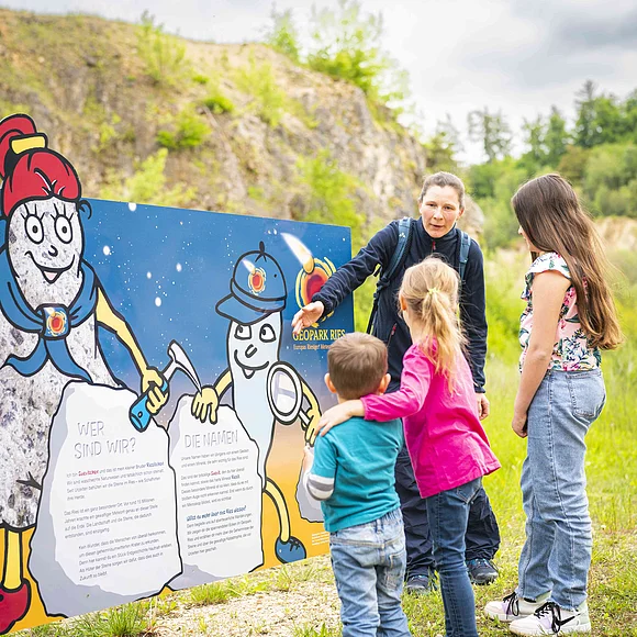 Eine Frau zeigt drei Kindern eine Informationstafel mit den Maskottchen Suevitchen und Riesitchen.