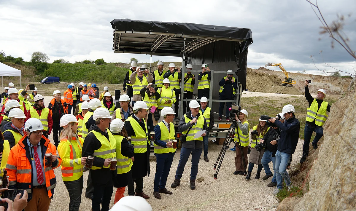 Eine große Gruppe Personen mit weißen Schutzhelmen und gelben Warnwesten steht mit Getränken in den Händen in einem Steinbruch.