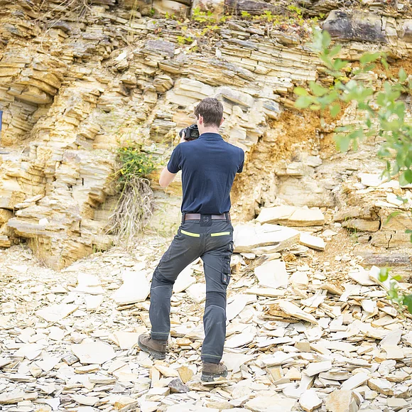 Ein Mann fotografiert mit einer Kamera eine Felswand aus plattigem Kalkgestein. Der Boden ist ebenfalls mit Bruchstücken von Kalkgesteinsplatten bedeckt.