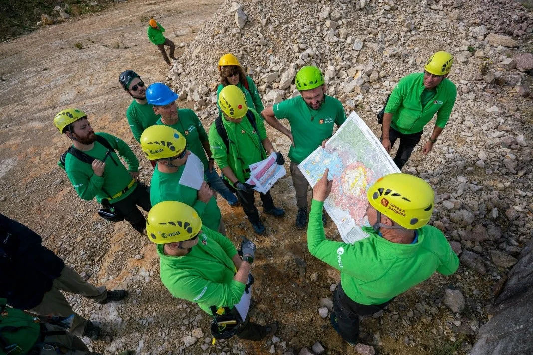Eine Gruppe Personen in grünen Oberteilen und mit Helmen steht in einem Steinbruch und betrachtet eine geologische Karte.