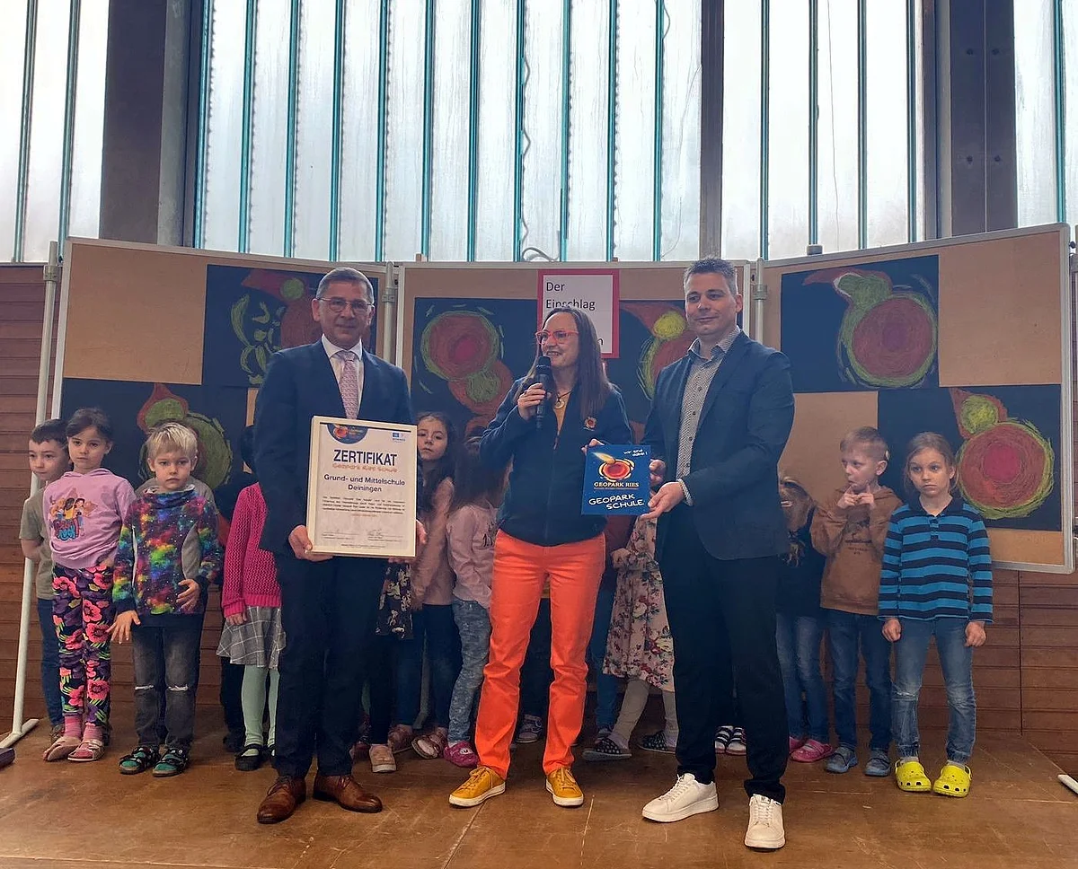Heike Burkhardt überreicht Wilhelm Rehklau und Andreas Langer die Urkunde der Geopark Ries Schule Deiningen. Dahinter stehen einige Kinder der Schule und gemalte Bilder des Meteoriteneinschlags.