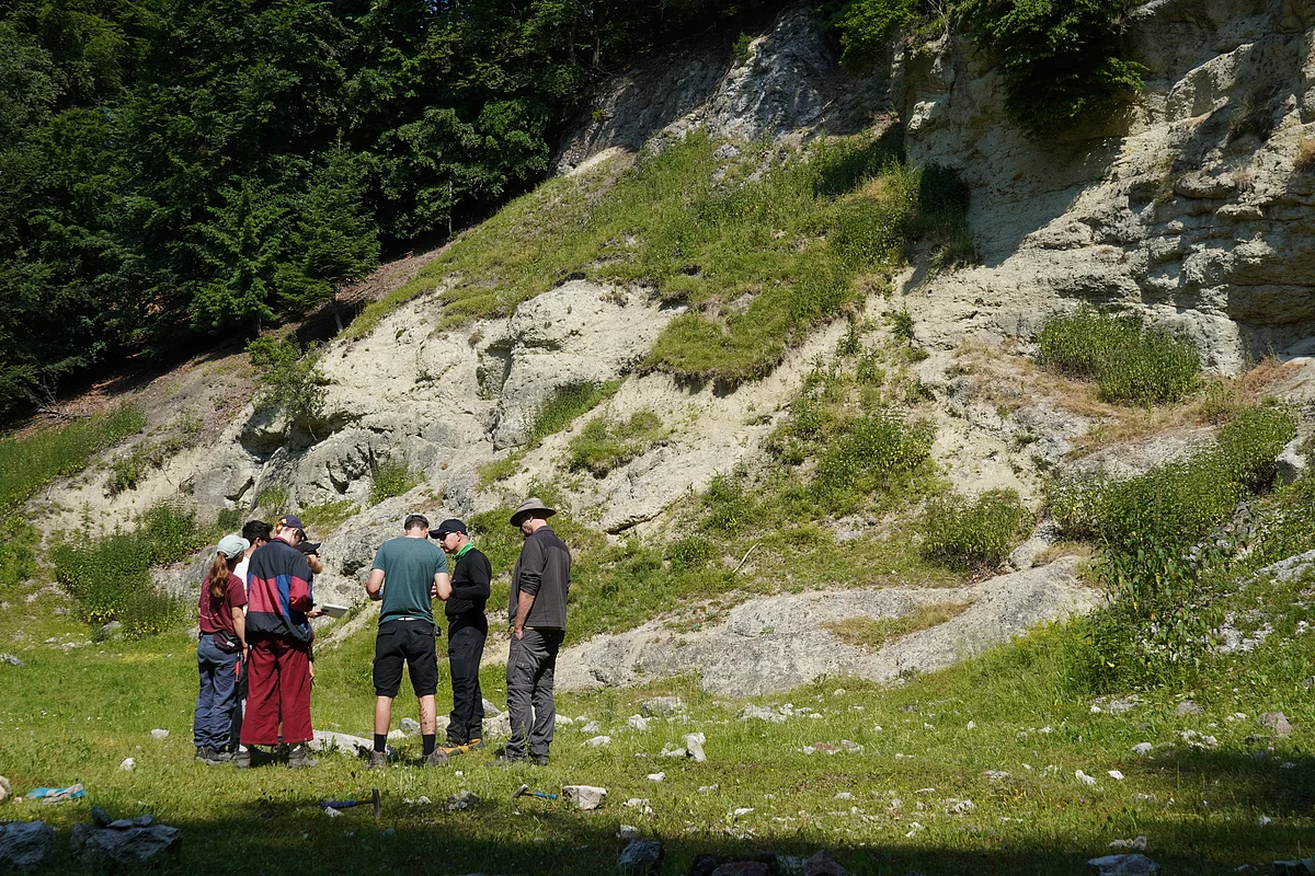 Eine Gruppe Personen steht vor einem Hang mit Felsaufschlüssen und Wiese.