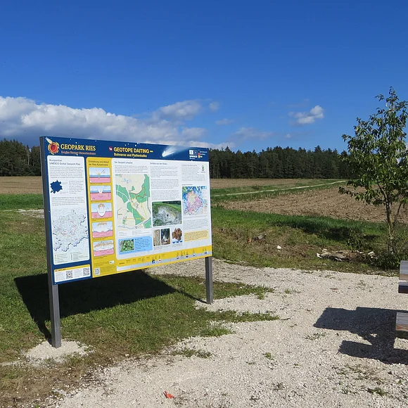 Auf dem Schotterplatz vor einer Informationstafel mit der Überschrift "Geotope Daiting" steht eine Sitzbank aus Holz.