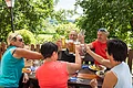 Eine Gruppe Personen sitzt um einen Holztisch in einem Biergarten und stößt mit Bierkrügen an.