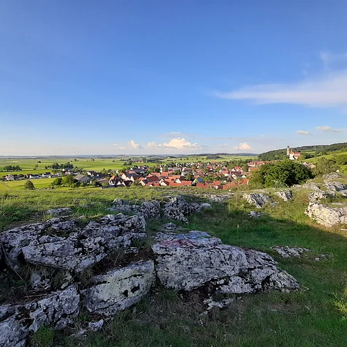 Von einem Hügel mit felsiger Kuppe aus erstreckt sich unter blauem Himmel der Blick in eine weite Landschaft mit einer Siedlung, Äckern, Wiesen und Bäumen.