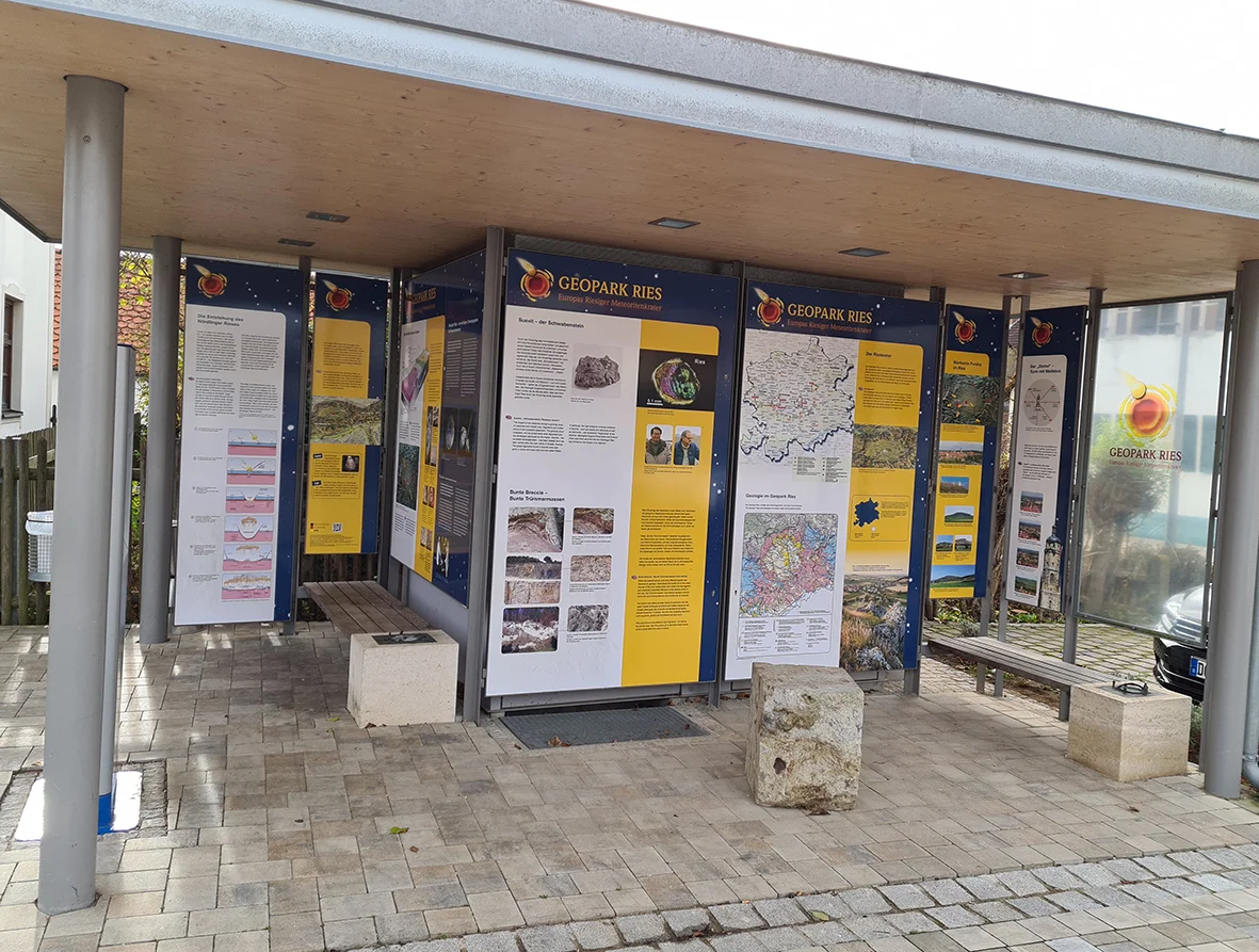 Unter einer Überdachung stehen mehrere Informationstafeln über den Geopark Ries und zwei Sitzbänke.