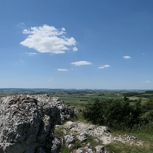 Hinter einem Felsblock erstreckt sich eine weite Landschaft. Am blauen Himmel befinden sich nur wenige weiße Wolken.