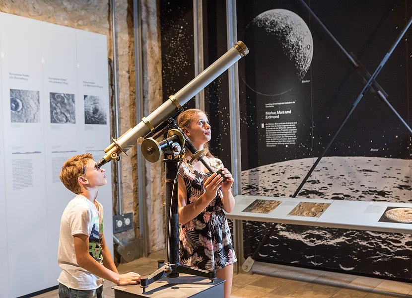 Ein Junge blickt durch ein Teleskop, neben dem ein Mädchen steht. Im Hintergrund sind große Plakate über Planeten zu sehen.