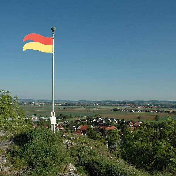 Von einem Felsen mit einer Metallfahne aus erstreckt sich der Blick über eine weite Landschaft mit Siedlungen, Äckern und Wald.