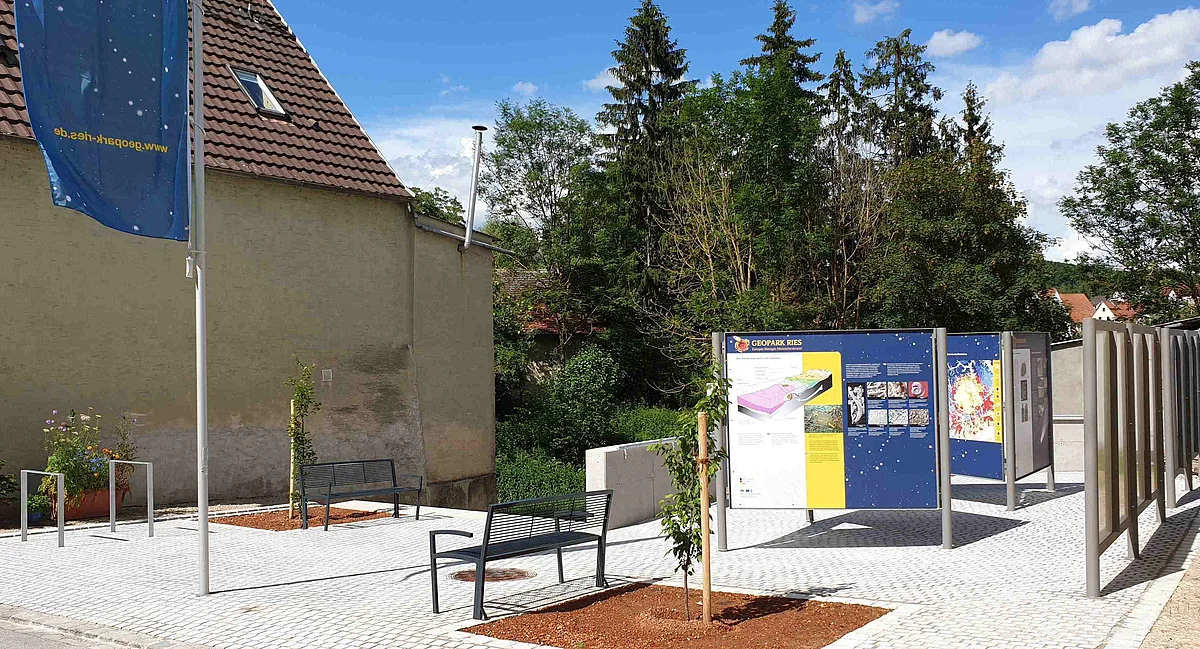 Auf einem gepflasterten Platz stehen mehrere Informationstafeln, Sitzbänke und zwei junge Bäume.