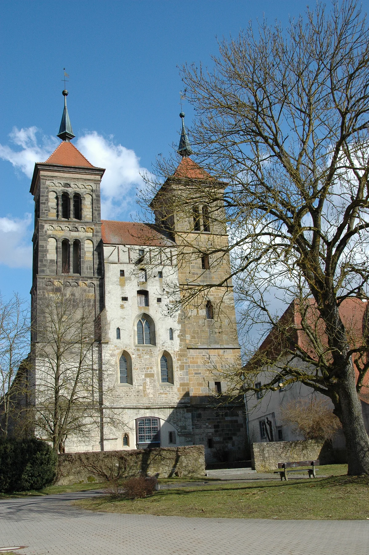 Zwischen zwei Türmen einer Kirche mit unverputztem Mauerwerk befindet sich eine hohe Stirnseite mit verschiedenartigen Fenstern.