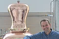 Ein Mann mit kariertem Hemd stützt seinen Ellbogen auf einen großen Kupferkessel.