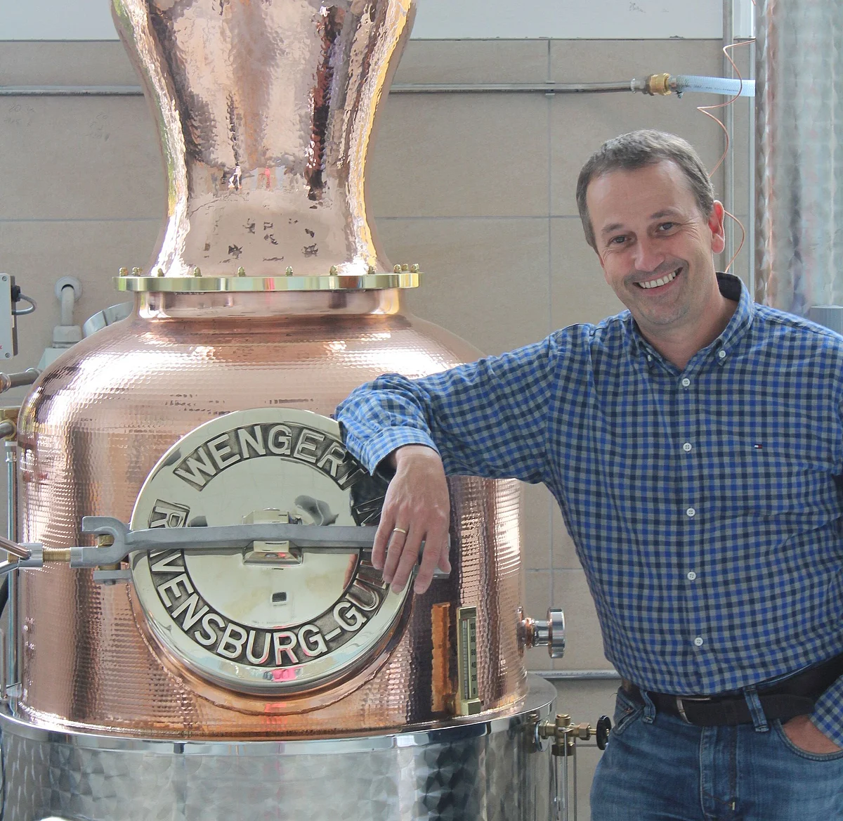 Ein Mann mit kariertem Hemd stützt seinen Ellbogen auf einen großen Kupferkessel.