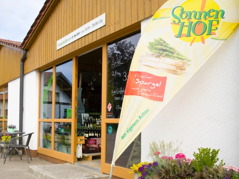 Durch eine geöffnete Glastür ist eine Palette mit Weinflaschen zu sehen. Neben der Tür steht eine Fahne mit der Aufschrift "Sonnenhof" und "frischer Spargel aus dem Donau-Ries - aus eigenem Anbau".
