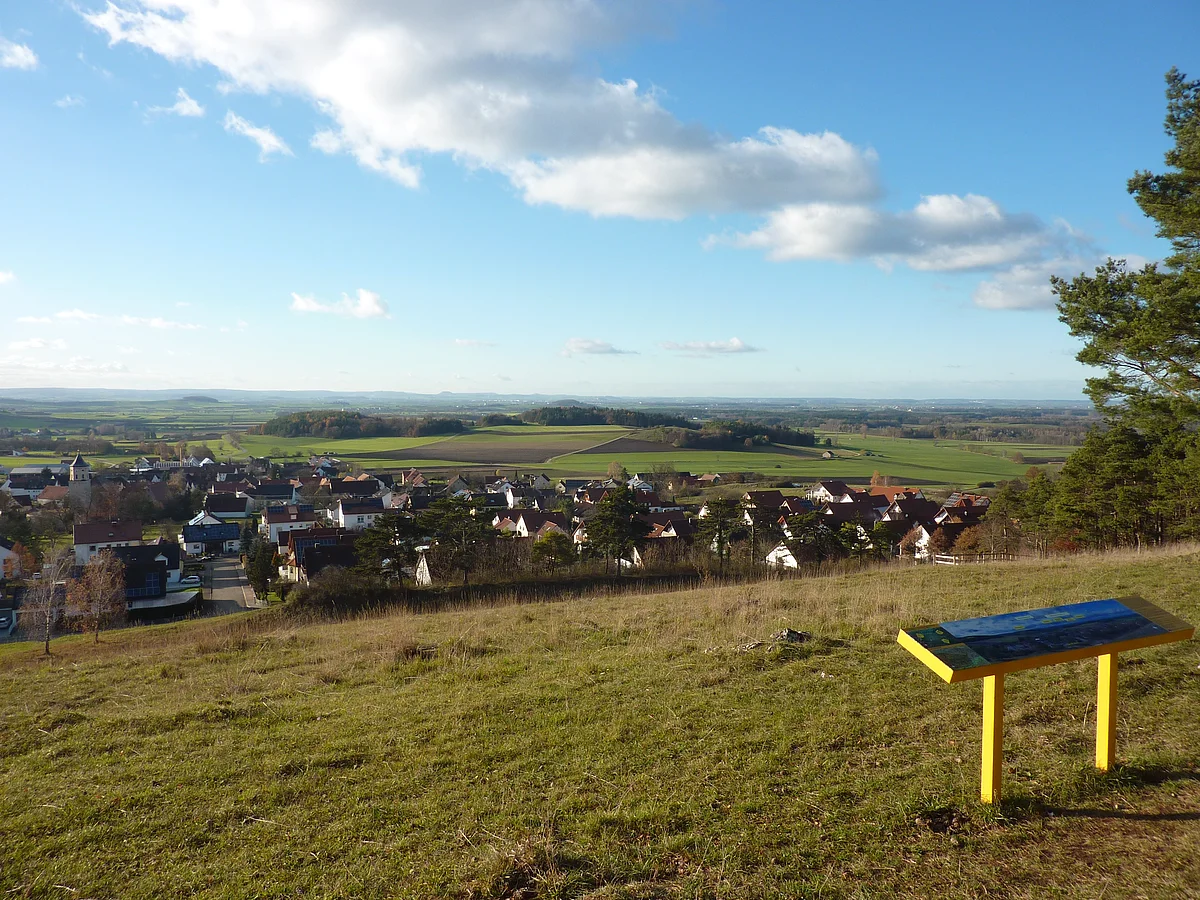 Von einem Hügel mit einer Panoramatafel aus erstreckt sich der Blick über eine Siedlung in eine weite Landschaft mit Äckern, Wiesen und Wald.