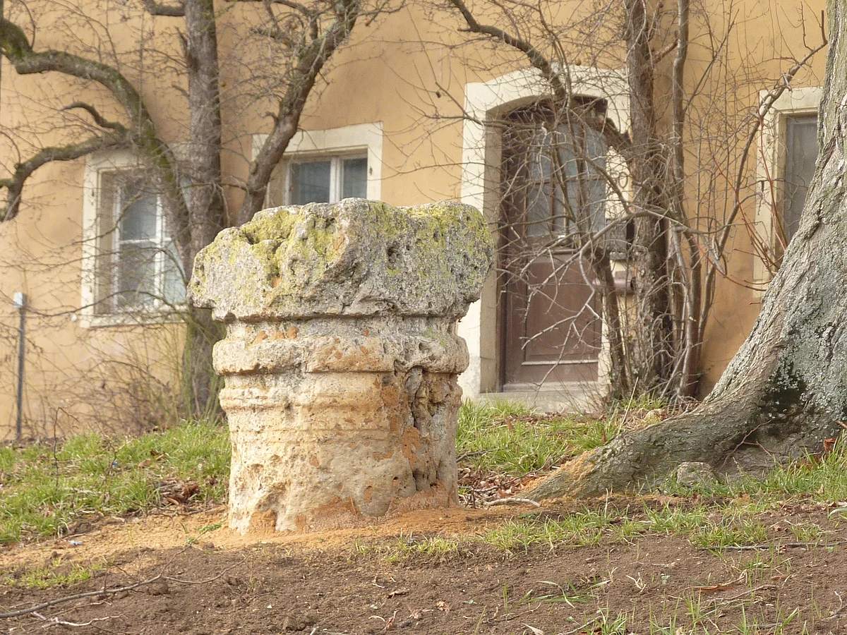 Ein säulenförmig behauener Stein steht neben einem alten Baum. Im Hintergrund ist eine ockerfarbene Hauswand zu sehen.