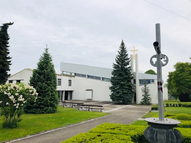 Blick auf die Kirche. Das Gotteshaus ist weiß, ein moderner Turm mit Kreuz, außen sind Bäume, Sträucher usw.