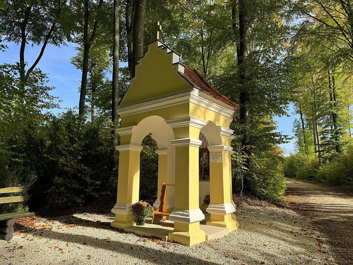 Eine gelbe kleine Kapelle steht an einem Waldweg. Außemherum sind grün belaubte Bäume.