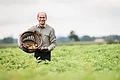 Ein Mann steht auf einem Feld mit hohen Kartoffelpflanzen und hält einen Korb mit einigen Kartoffeln in den Händen.