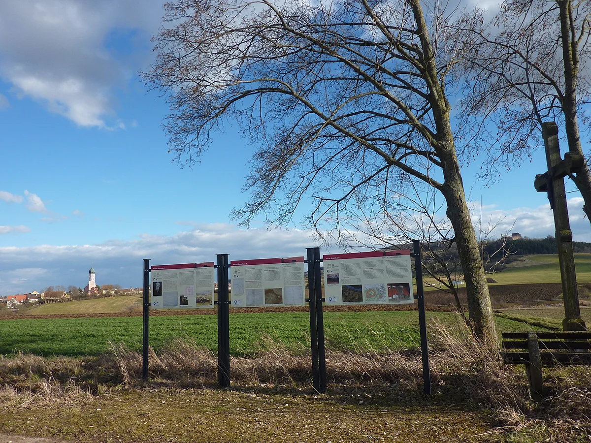 Am Rande eines Ackers stehen drei Informationstafeln neben zwei Bäumen und einem Wegkreuz. In der Ferne ist eine Siedlung mit Kirchturm zu sehen.