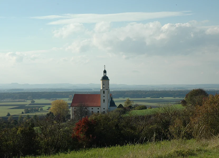 Eine Kirche mit weiß-gelber Fassade steht leicht erhöht. Im Hintergrund erstreckt sich eine weite Landschaft.