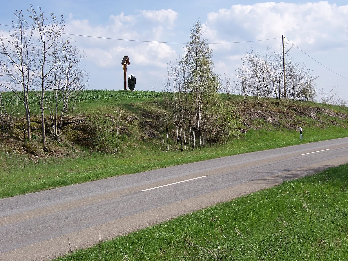 An der Böschung neben einer Straße sind mehrere Felsen und kleine Bäume zu erkennen. Auf der Böschung steht ein Wegkreuz.