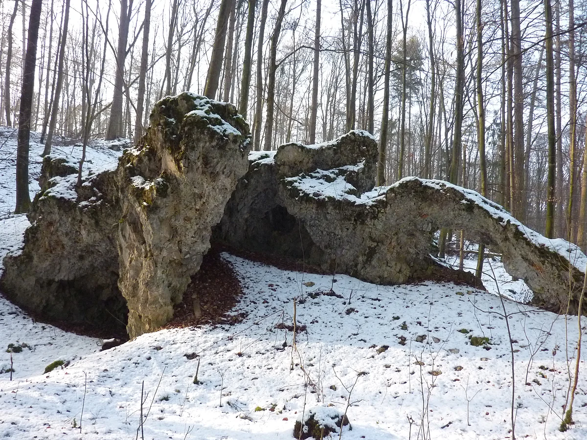 In einem Wald befindet sich eine mehrteilige Felsformation mit einem kleinen Bogen. Auf dem Boden und auf den Felsen liegt Schnee.