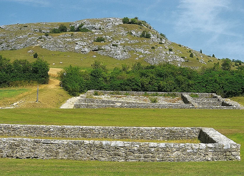 In einer Wiese befinden sich Mauerfundamente zweier Gebäude. Hinter einer Buschreihe erhebt sich ein felsiger Hügel.