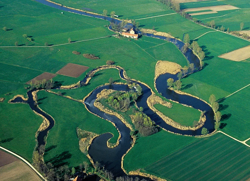 In der Nähe eines Gehöfts mäandriert ein Fluss umgeben von Wiesen und Äckern.
