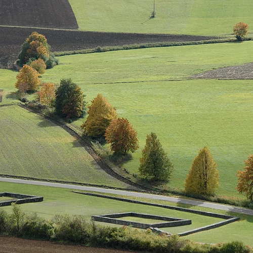 Neben einem schmalen Weg befinden sich Mauerfundamente in einer Wiese. Auf der anderen Wegseite verläuft eine Baumreihe zwischen Wiesenstücken und Äckern.