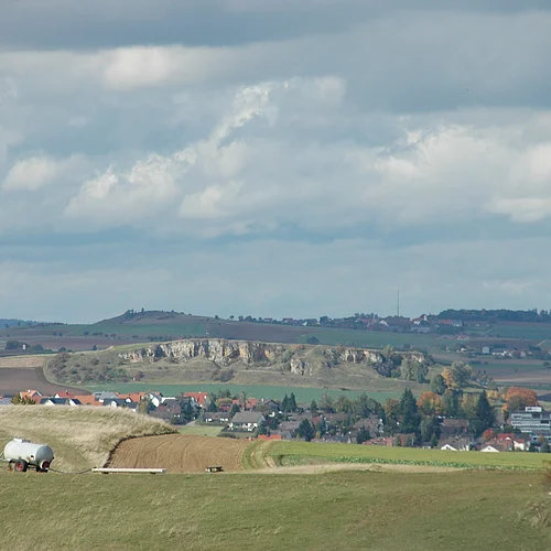 Hinter einer Siedlung in einer Senke erhebt sich ein Hügel mit felsiger Front. Am Horizont sind weitere Hügel mit Wald zu sehen.