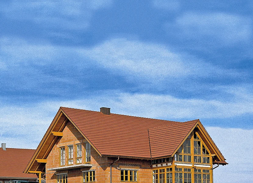 Auf einer leichten Anhöhe steht ein unverputztes Ziegelhaus mit einem Anbau mit vielen Fenstern.