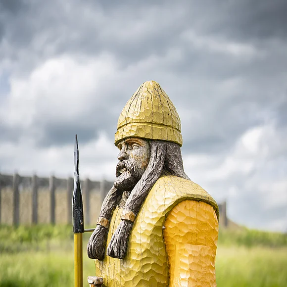 Geopark-Ries Juni 2022 Auf einer Wiese steht die aus Holz geschnitzte und bemalte Figur eines bewaffneten keltischen Mannes. Im Hintergrund ist ein rekonstruiertes Bauwerk zu sehen.