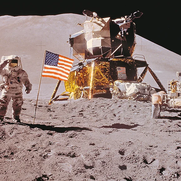 Ein Astronaut steht neben einer USA-Flagge auf der Mondoberfläche. Im Hintergrund sind zwei Mondrover zu sehen.