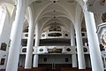 Innenansicht einer Kirche, der Stadtpfarrkirche St. Walburga in Monheim, mit hohen weißen Säulen, Gewölbedecke, Emporen mit Verzierungen und Holzbänken.