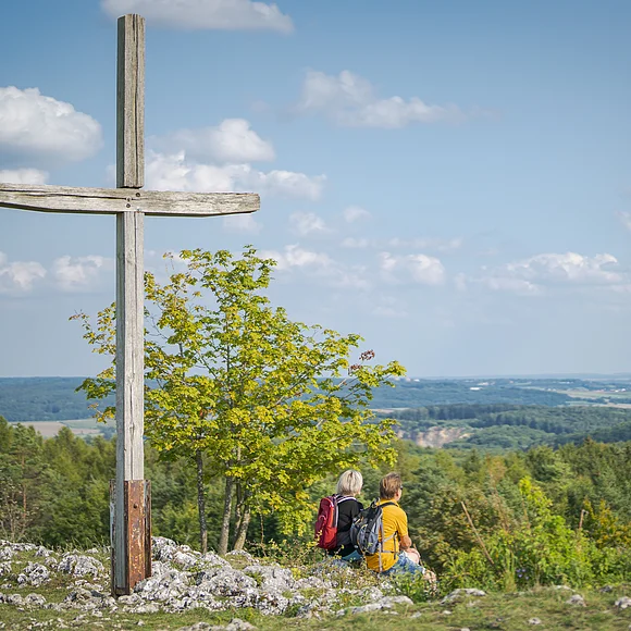 Holzkreuz auf dem Harburger Bockberg, zwei Personen mit Rucksäcken sitzen daneben, Blick auf die bewaldete Landschaft.