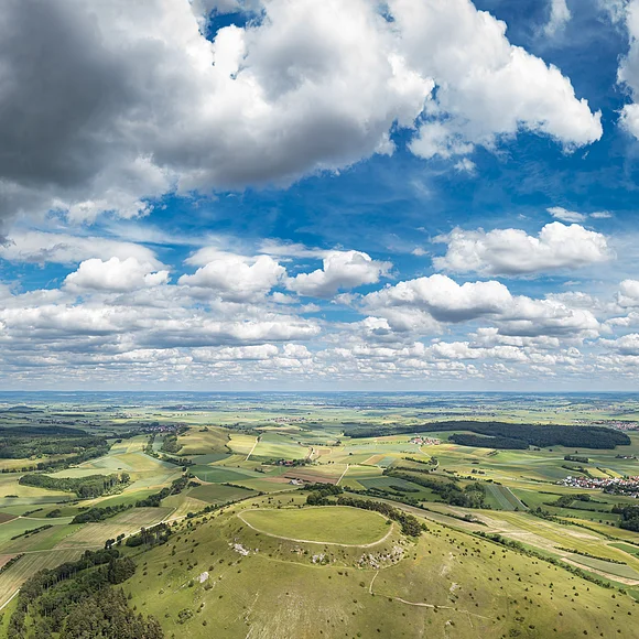 Weite Landschaft mit Feldern, Hügeln und einem Dorf unter einem Himmel mit vielen Wolken.