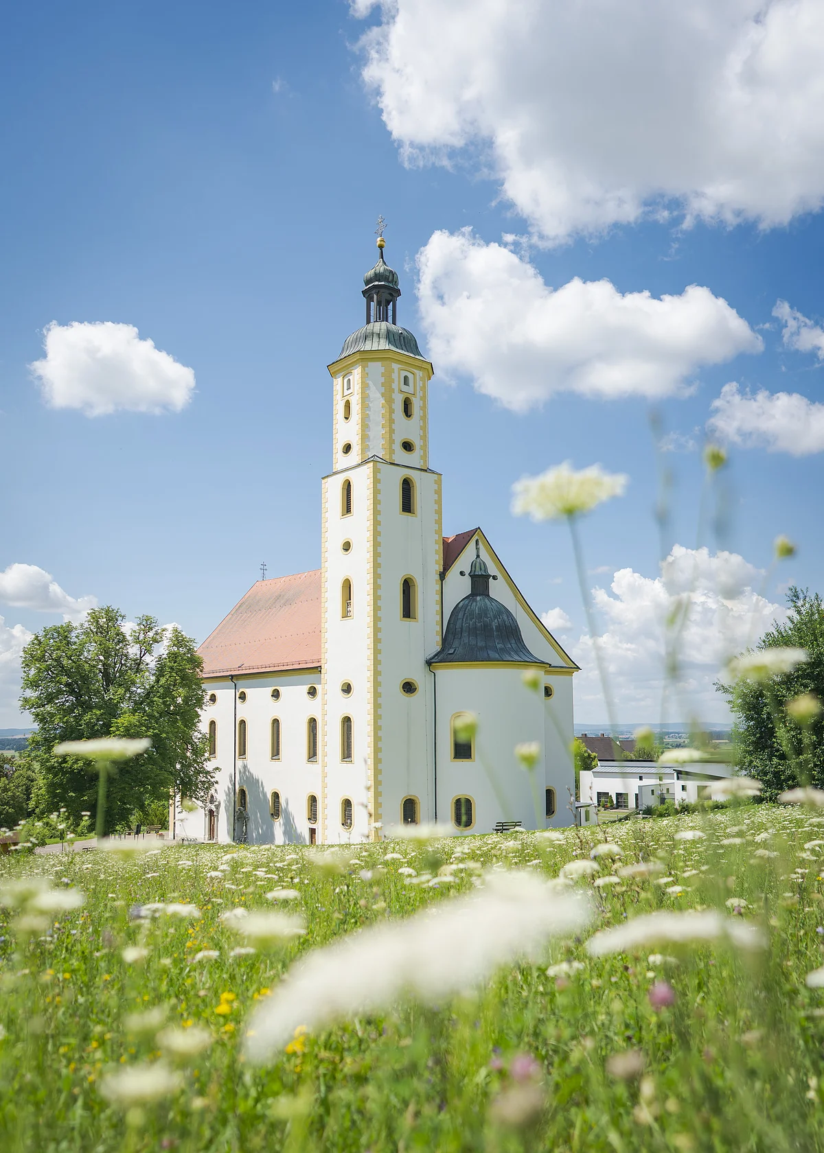Kirche mit Turm und rotem Dach auf einer Wiese, blauer Himmel mit Wolken im Hintergrund.