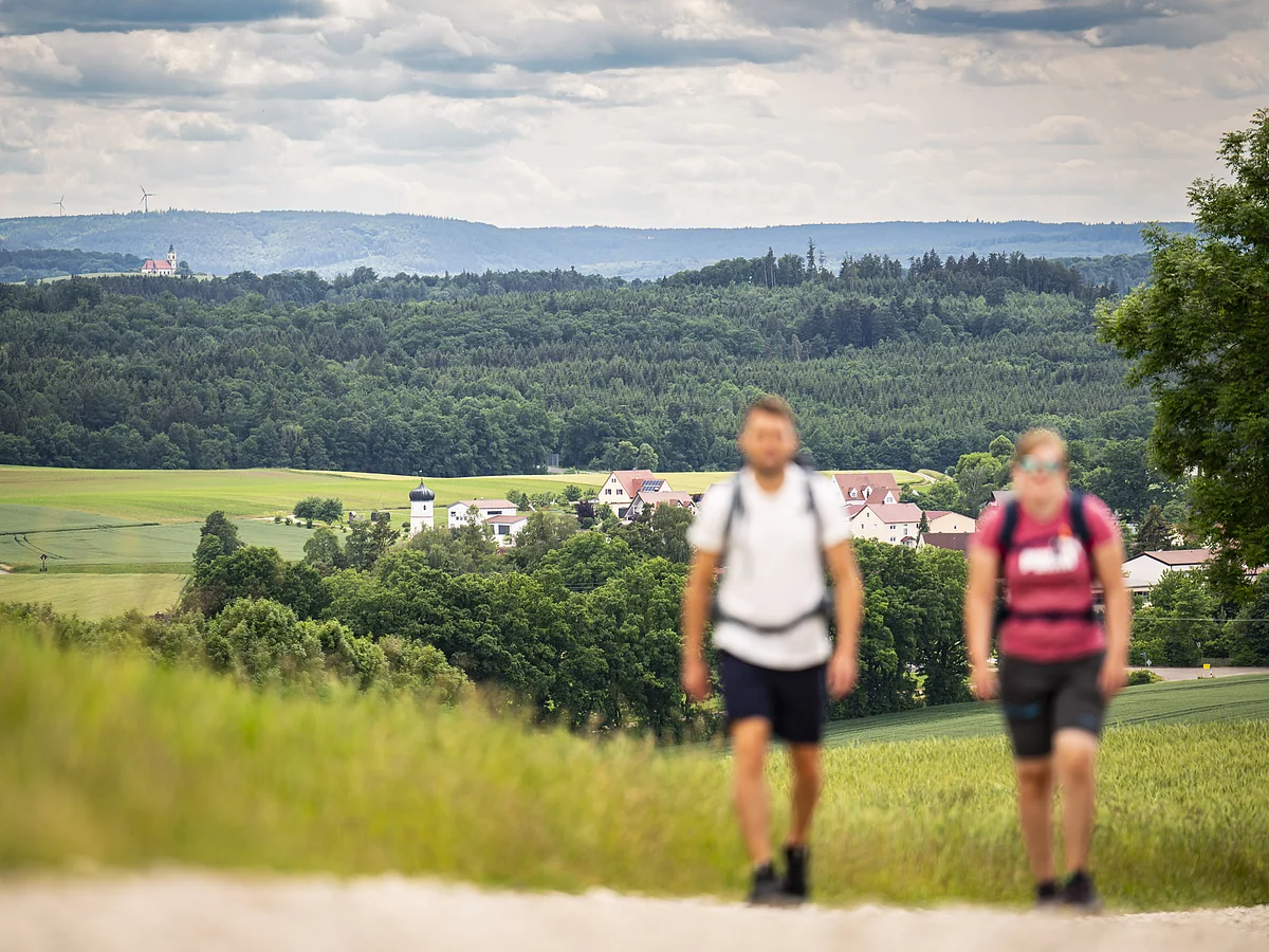 Zwei Wanderer an der Weidenkapelle bei Fremdingen Zwei unscharfe Personen wandern auf einem Weg, im Hintergrund eine Landschaft mit Feldern, Wald und Gebäuden.