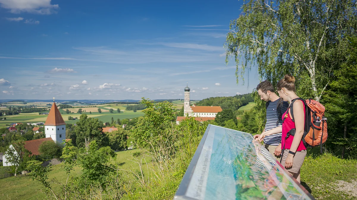 Zwei Personen betrachten eine Informationstafel mit Blick auf eine ländliche Landschaft mit Kirche und Kloster.