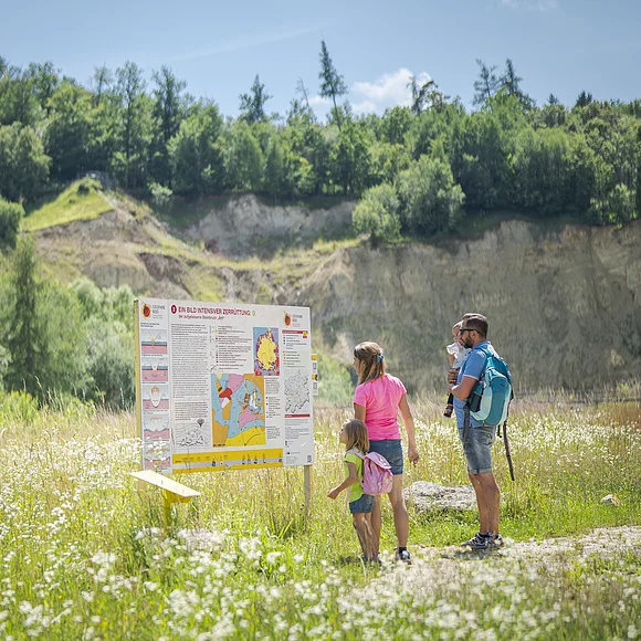 Eine Familie steht vor einem Informationsschild in einer grünen Landschaft mit Hügeln im Hintergrund.