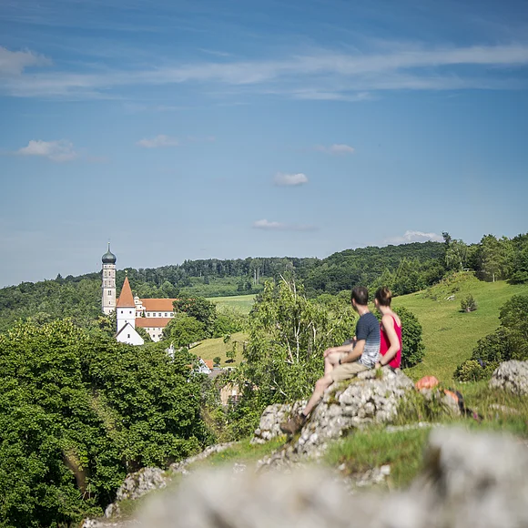Zwei Personen sitzen auf einem Felsen und blicken auf eine Kirche in einer grünen Landschaft.