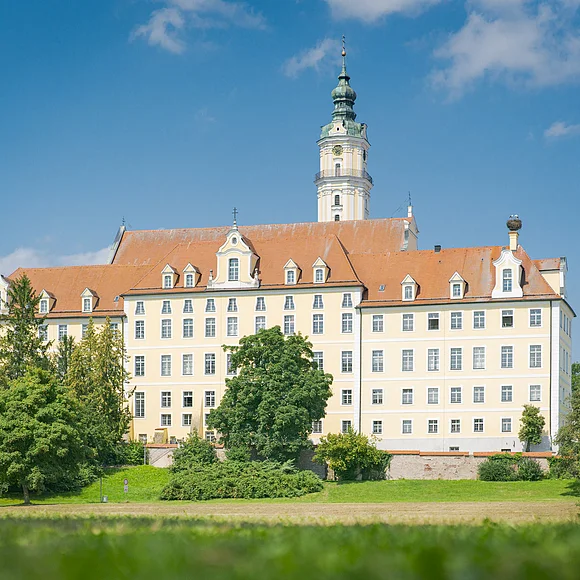 Großes, mehrstöckiges Gebäude mit orangefarbenem Dach und Turm, umgeben von Bäumen und Wiese, unter blauem Himmel.