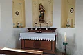 Kleiner Kirchenaltar einer Kapelle mit Marienstatue, Kerzen und Blumenschmuck vor weiß-gelb gestrichener Wand mit Ausfräsungen in der Wand mit gemalten Verzierungen darin. Im Vordergrund eine Gebetsbank aus Holz.