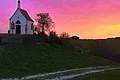 Links im Bild eine kleine Kapelle auf einem Hügel mit einem Weg unten am Hügel, umgeben von Bäumen und Weide. Sonnenuntergang mit pink-orangefarbenem Himmel im Hintergrund.