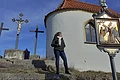 Frau steht auf einer Steintreppe neben einer Kapelle mit drei Kreuzen im Hintergrund und einem religiösem Bild auf einer Tafel, bei blauem Himmel.