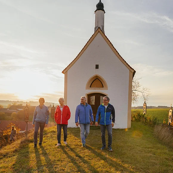 Kalvarienbergkapelle "Zur schmerzhaften Muttergottes" - umgeben von 14 Kreuzwegstationen in Wittesheim Vier Personen gehen nebeneinander auf einer Wiese vor einer kleinen Kapelle im Sonnenuntergang spazieren. Im Hintergrund grüne Wiese und weitere Gebäude.
