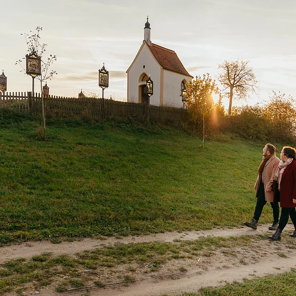Die Kalvarienbergkapelle "Zur schmerzhaften Muttergottes" in Wittesheim - eingerahmt von 14 Kreuzwegstationen Vier Personen spazieren auf einem Weg an einer kleinen Kapelle vorbei, der Kalvarienbergkapelle in Wittesheim, umgeben von herbstlicher Landschaft. Auf dem Weg vor der Kapelle stehen Kreuzwegstationen.