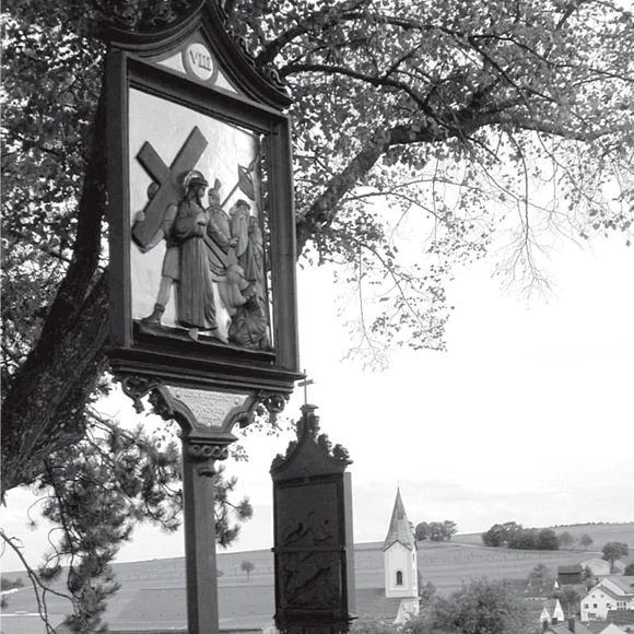 Kreuzweg-Station am Kalvarienberg in Wittesheim Schwarz-weiß Foto der Kreuzweg-Station am Kalvarienberg in Wittesheim mit einem Wegkreuz mit einer Kreuzigungsszene und dem Dorf mit Kirche im Hintergrund, unter Baumzweigen bei Tageslicht.